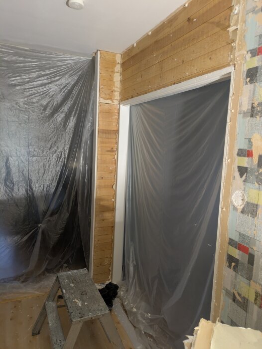 Rivningsprojekt med gipsborttagning; stege, plastskydd, avtäckta träväggar och synliga gipsskivor i inomhusmiljö.