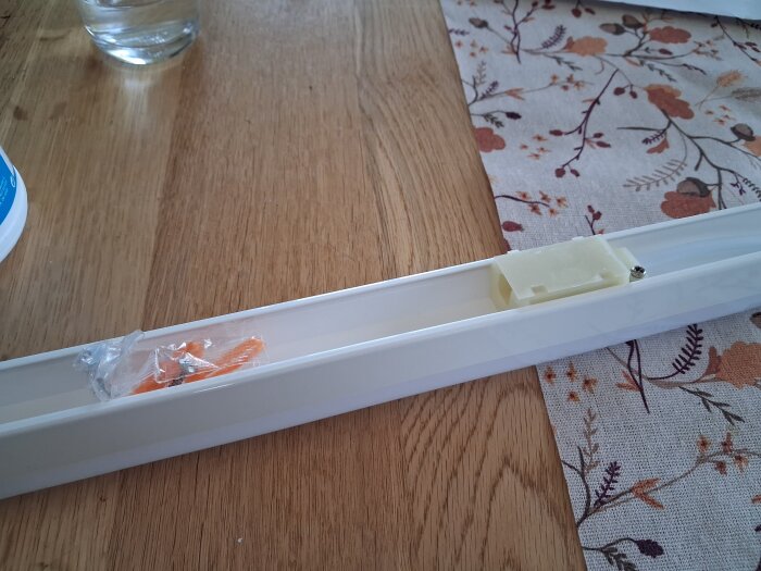 Vit lysrörsarmatur med monteringsdetaljer på ett träbord, intill en mönstrad duk och ett glas vatten.