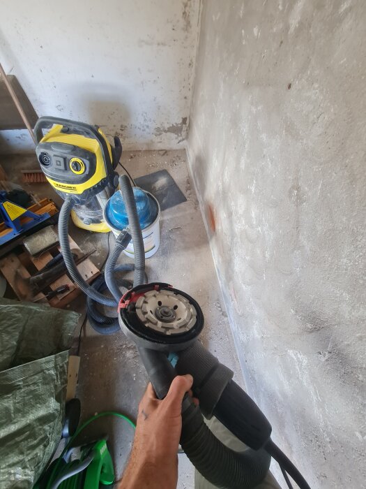 Hand håller en vinkelslip med diamantkopp i ett rum med avskavd puts på väggen och en dammsugare i bakgrunden.