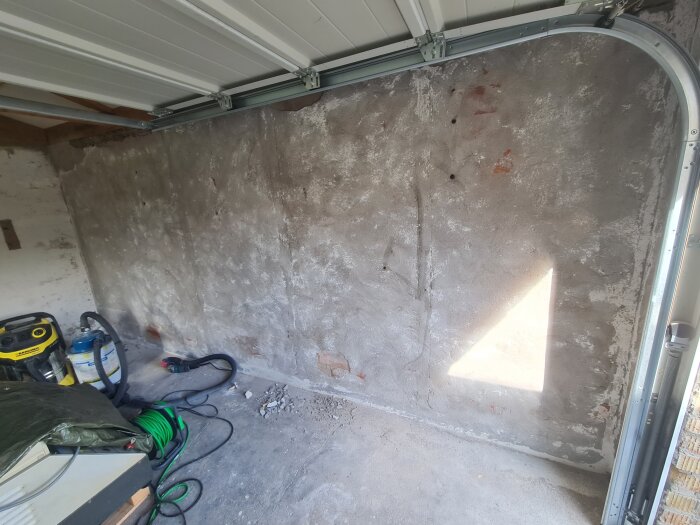Vägg i garage med avskavd puts, delvis solbelyst. Verktyg, inklusive vinkelslip, och kablar syns på golvet intill.
