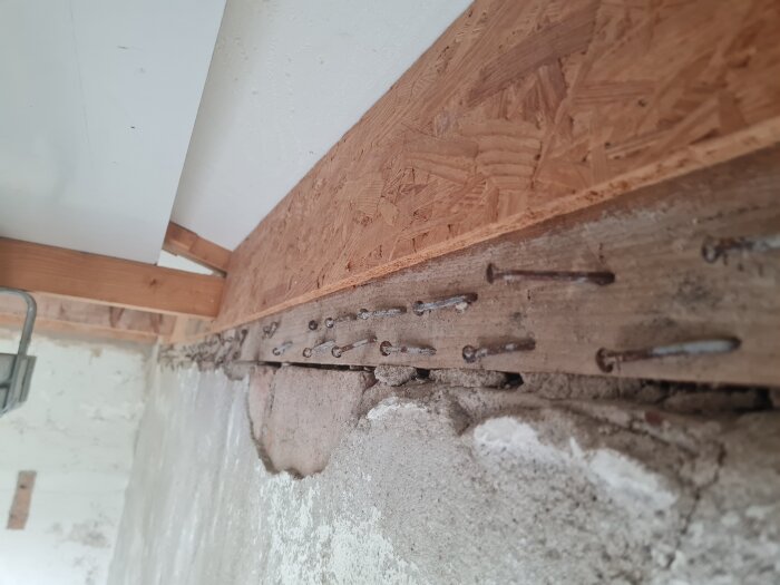 Träregel med flera spikar och exponerad betongvägg visar sprickor och gammal puts.