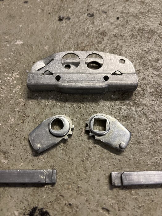 Mekaniska delar, inklusive en större metallbit med hål ovanför två mindre kugghjulsliknande objekt, placerade på en betongyta.
