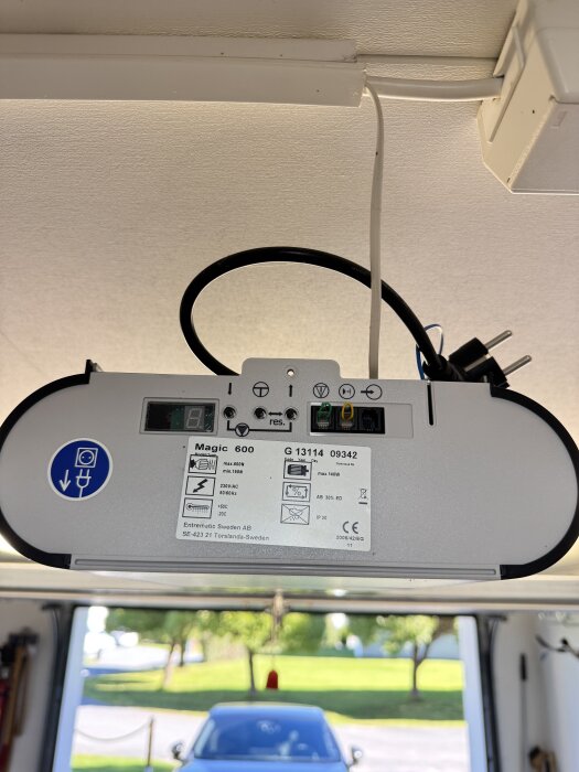 Motor Magic 600 med borttagen front täcker en garageöppnare. Sökande efter skruv för demontering. Bakgrund visar bil och grönskande utomhus.