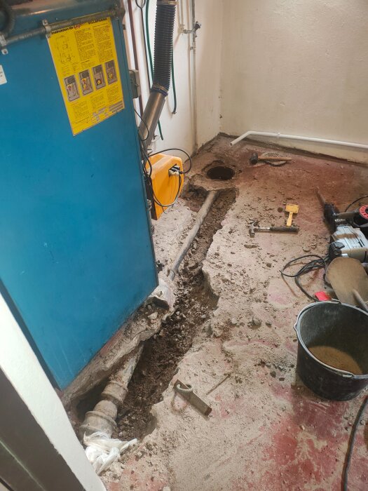 Byggarbetsområde med blottat rör i golvet nära en blå panna, omgiven av verktyg och en hink. Hål i golvet visar rörinstallation.