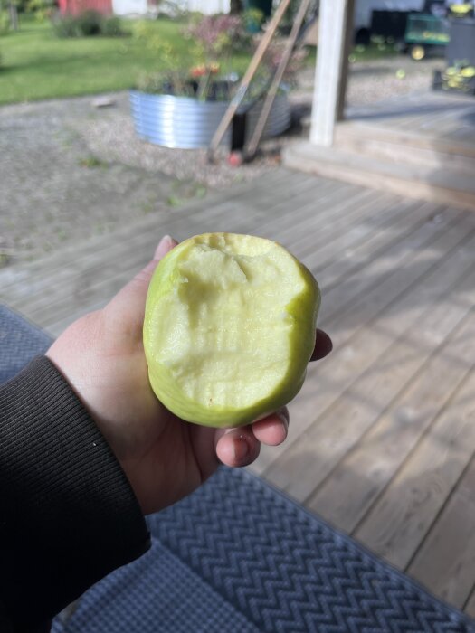 En hand håller ett grönt äpple med en tugga tagen, på en träaltan med trädgård i bakgrunden.