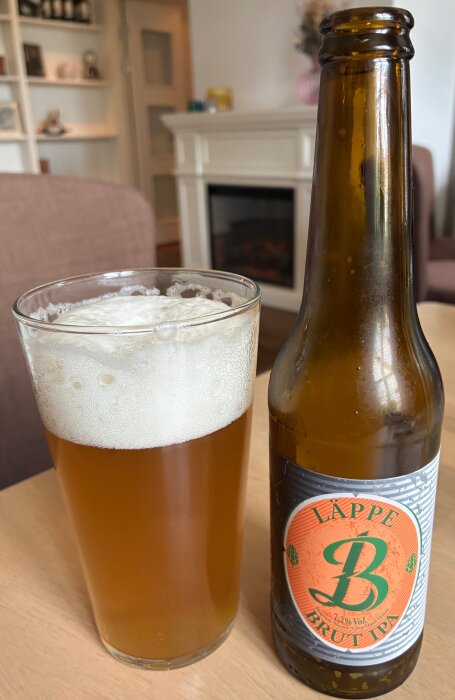 Ölglas med skum vid sidan av en flaska med etikett där det står "Läppe Brut IPA", på ett träbord i ett vardagsrum.