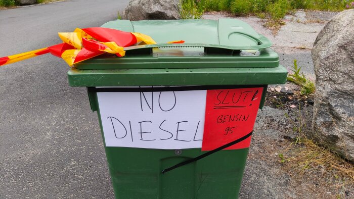 Grön soptunna med skyltar som säger "NO DIESEL" och "SLUT! Bensín 95", samt en röd och gul vimpel ovanpå, på asfaltväg.