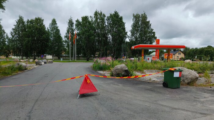 Avspärrad väg vid bensinstation med träd i bakgrunden. En grön soptunna och en röd vägskylt med gulröd tejp stänger av vägen.