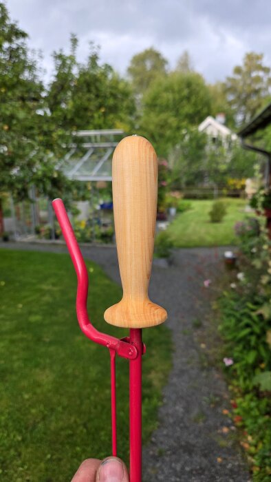 Röd griptång med nyss svarvat handtag i trä, fotograferad i en frodig trädgårdsmiljö.