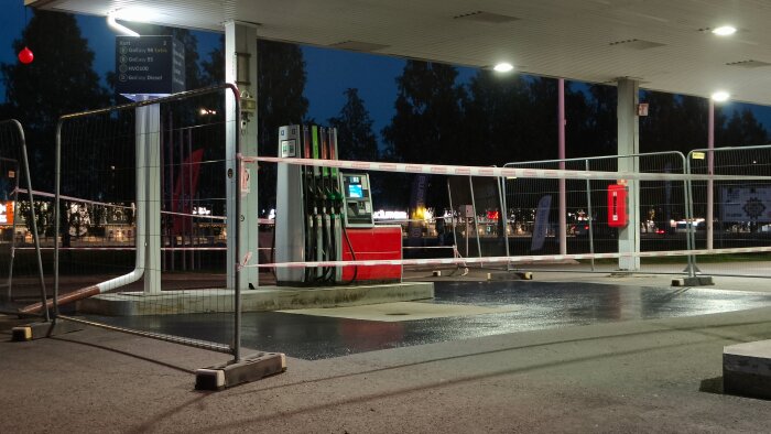 Avspärrad bensinstation med byggstängsel. Fyra bränslepumpar synliga. Night time-setup under upplyst tak.