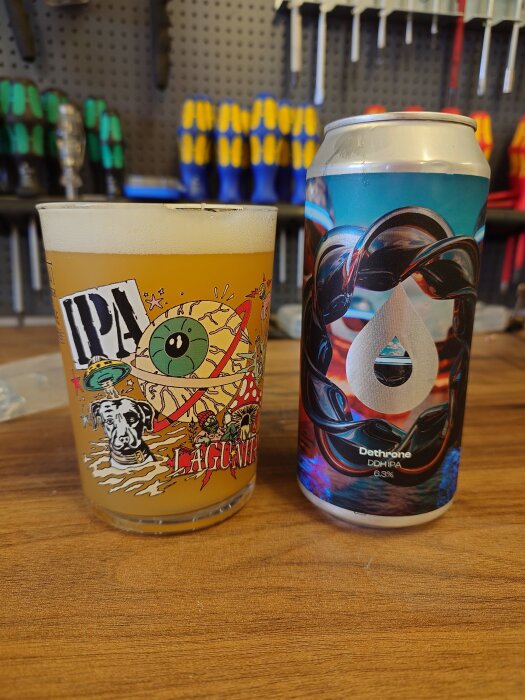 En glas IPA och en ölburk med färgglad etikett står på ett träbord i bakgrunden av verktyg.