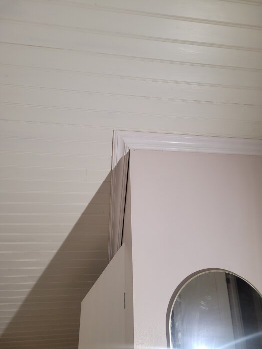 Innertak i panel med snedtak och en lös taklist ovanför en vägg med ett runt spegelutrymme.