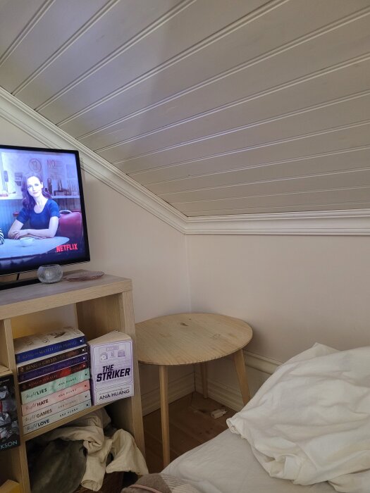 Snedtak och vägg med lister, TV på hylla med böcker, runt bord och säng i ett sovrum.