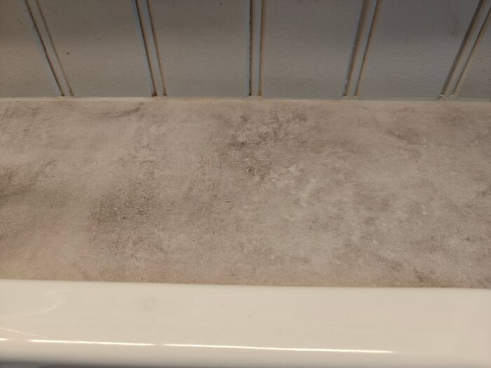 Väggpanel med pärlspont ovanför ett golv med betongliknande yta, fogad med silikon vid basen. Grov textur synlig på ytan.