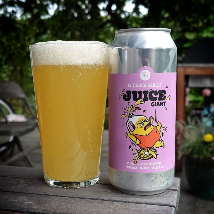 Ölburk med etiketten "Other Half Juice Giant" och ett glas skummande öl står på ett trädäck, omgiven av grönska.