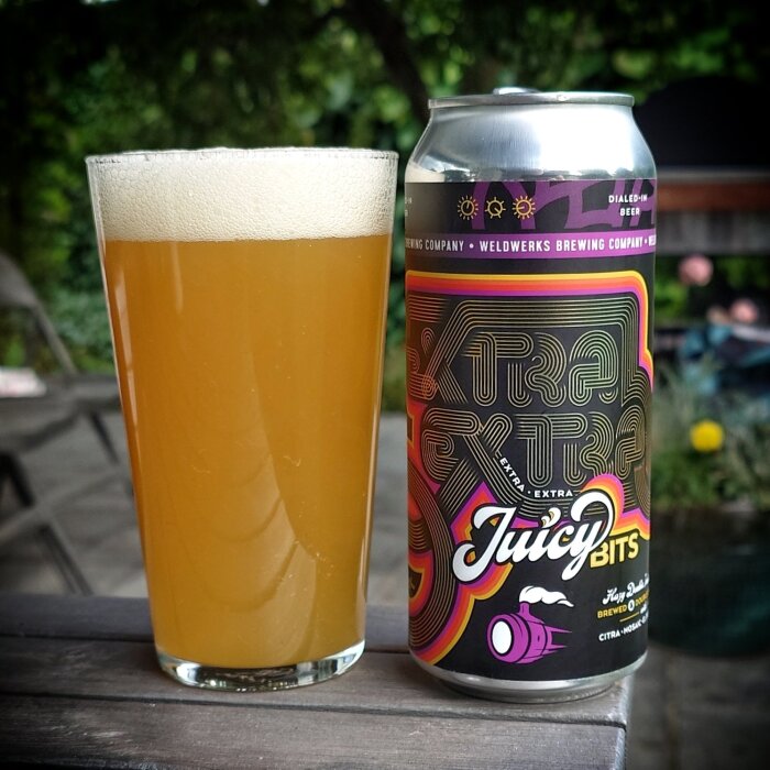 En burk Weldwerks "Juicy Bits" öl bredvid ett glas med skummande, gyllene öl på ett utomhusbord i grönska.