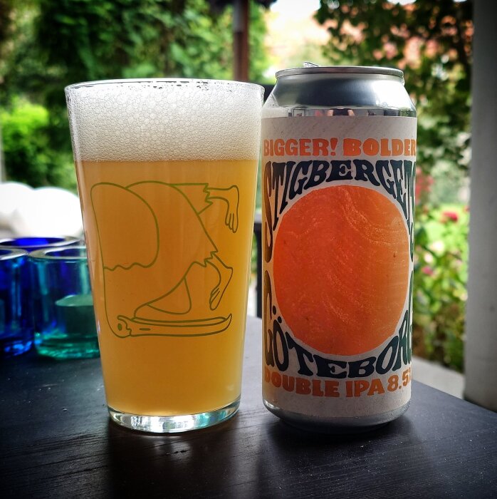 Ett glas hazy dubbel IPA bredvid en burk Stigbergets öl på ett bord utomhus, omgiven av grönska.