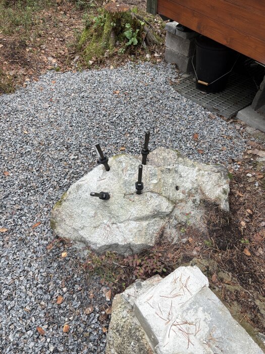 Fem HME-verktyg på en sten omgivna av grus och naturlig vegetation vid en byggnad.