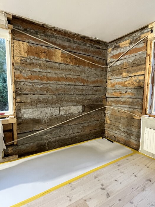 Timmervägg under renovering i vardagsrum med exponerat trä och drevade springor, förberedd för lerklining. Fönster syns till vänster.