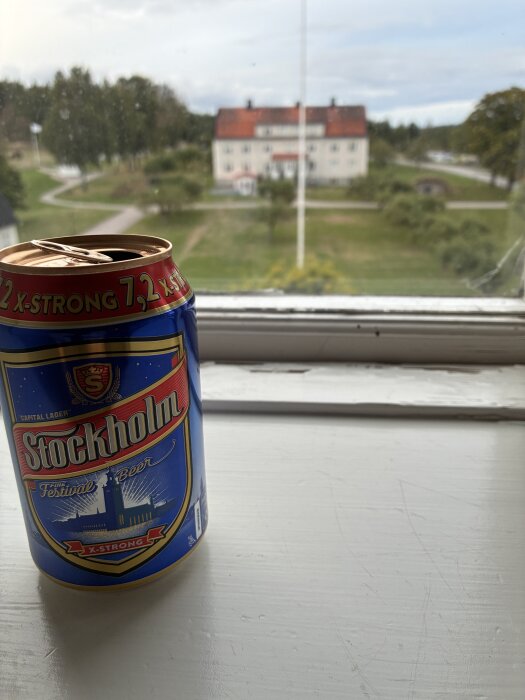 Blå ölburk med texten "Stockholm" på fönsterbräda, utsikt över en stor vit byggnad och grönska utanför.
