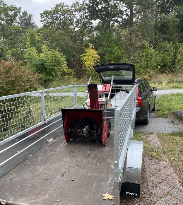 Bil med släpvagn parkerad på uppfart, lastad med en röd snöslunga framför öppet bagageutrymme, omgiven av grönskande träd och buskar.