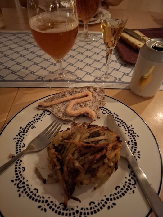 Tallrik med potatisrätt och knäckebröd med ost, ölglas, snapsglas och konservburk med fågelmotiv på bord.