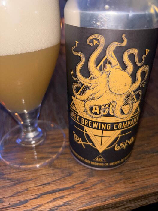 En burk Apex Brewing Company IPA med en bläckfiskillustration och ett glas öl med skumkrona på ett trägolv.