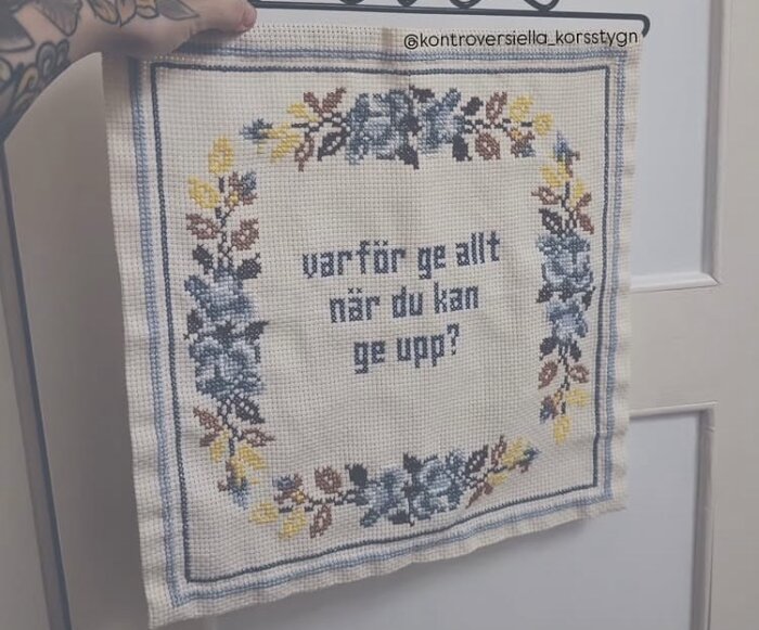 Broderad bonad med texten "varför ge allt när du kan ge upp?" omgiven av blommor i blått, gult och brunt, hålls upp mot en dörr.