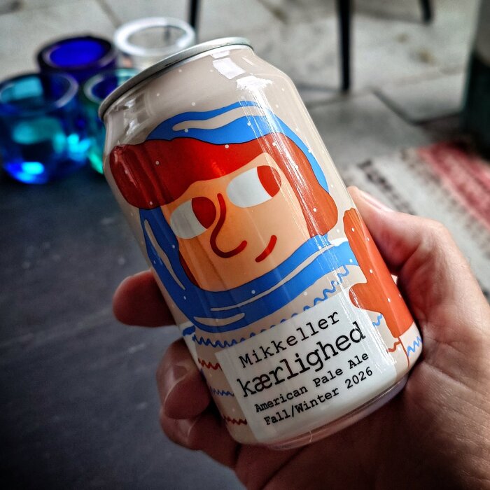 En hand håller en Mikkeller Kærlighed American Pale Ale-burk med färgglad illustration.
