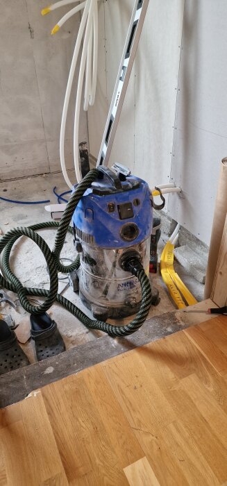 En blå dammsugare av märket Nilfisk står i ett rum under renovering, ansluten med en grön-randig slang.