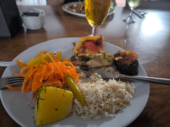 Tallrik med ris, grönsaker, någon typ av kycklingrätt och bröd. En öl och ett glas vin i bakgrunden på en träbord.