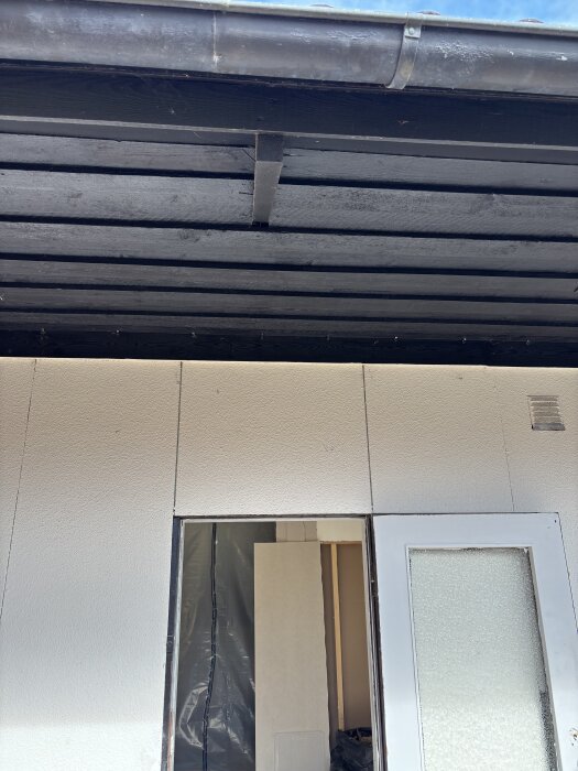 Fasad av hus med en delvis borttagen dörrkarm och svartmålat tak ovanför; renovering pågår med dörrbyte och ombyggnation.