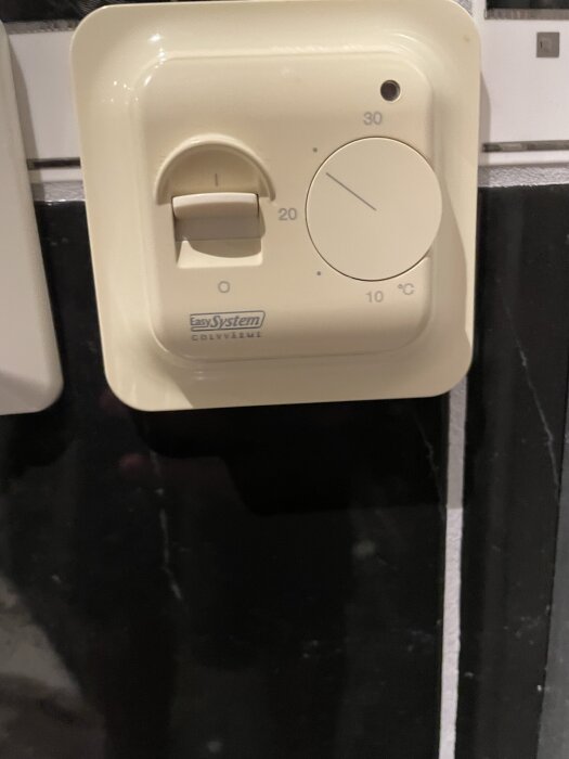 Bilden visar en beige golvvärmetermostat av märket "Easy System" med en ratt som visar temperaturinställning från 10 till 30 grader Celsius.
