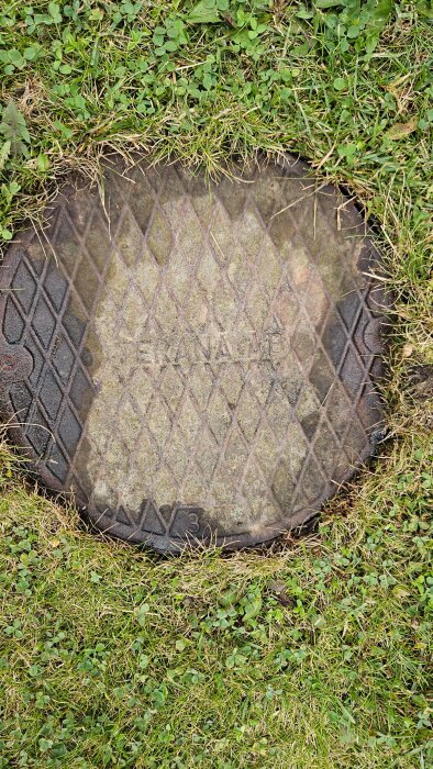 Brunnlock i metall med rutmönster, delvis täckt av mossa, omgivet av gräs. Text på locket: "Tekana AB".