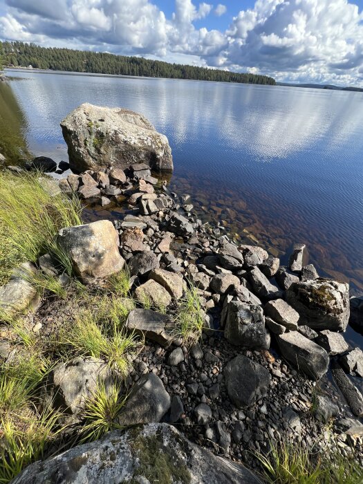 Stenig strandkant vid en sjö med blått vatten och grön skog i bakgrunden under en delvis molnig himmel.