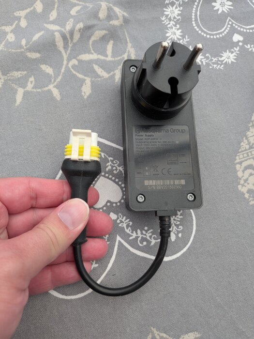 Hand håller en svart nätadapter från Husqvarna med en EU-kontakt. Adapterinformationen syns på etiketten. Ligger på grått mönstrat tygunderlag.