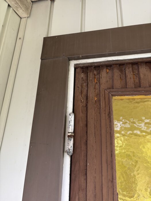Dörr med rostig gångjärn, brun karm, vit ram, och gul mönstrad glas, mot vit och brun panelvägg.