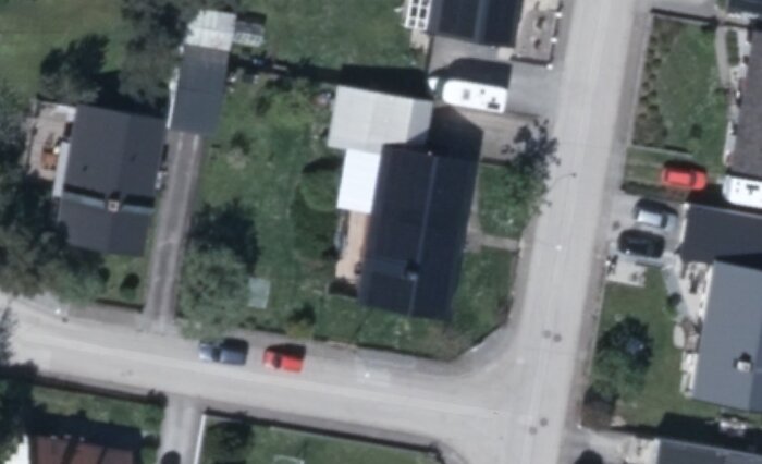 Satellitbild över hus och trädgård på hörntomt med intilliggande vägar, gröna områden och parkerade bilar.