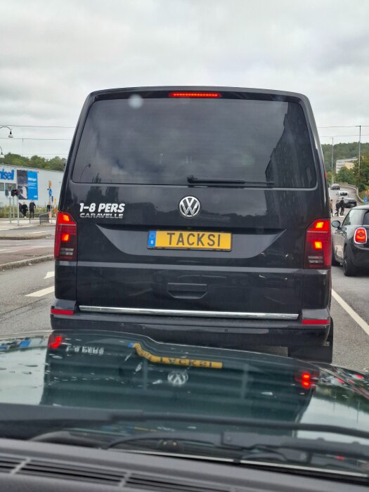 Svart Volkswagen Caravelle med registreringsskylten "TACKSI" syns bakifrån, står på en väg med andra bilar i bakgrunden.