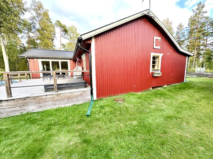 Rött trähus från 1930-talet med träfönster och veranda, omgiven av gräsmatta och skog. Visar utsidan och trädgården.