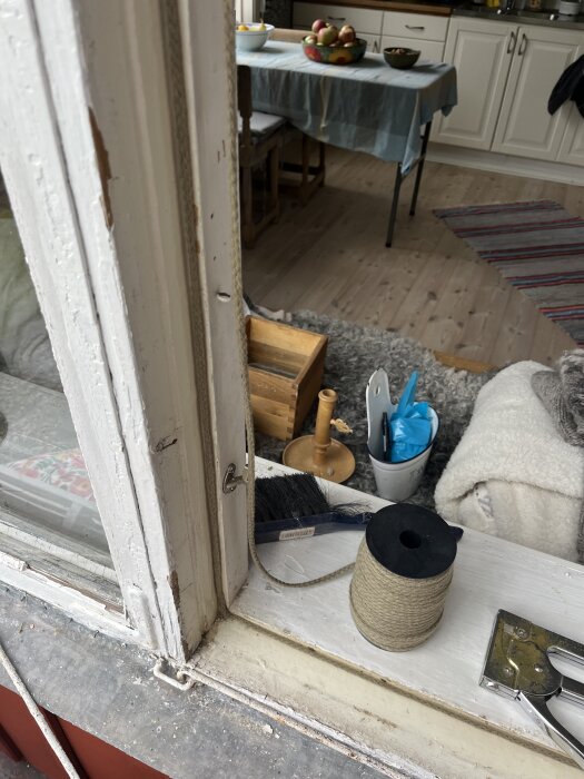 Fönsterkarm av trä med ull-snöre, pensel och häftpistol på fönsterbrädan. Bord med fruktskål synligt i bakgrunden inomhus.