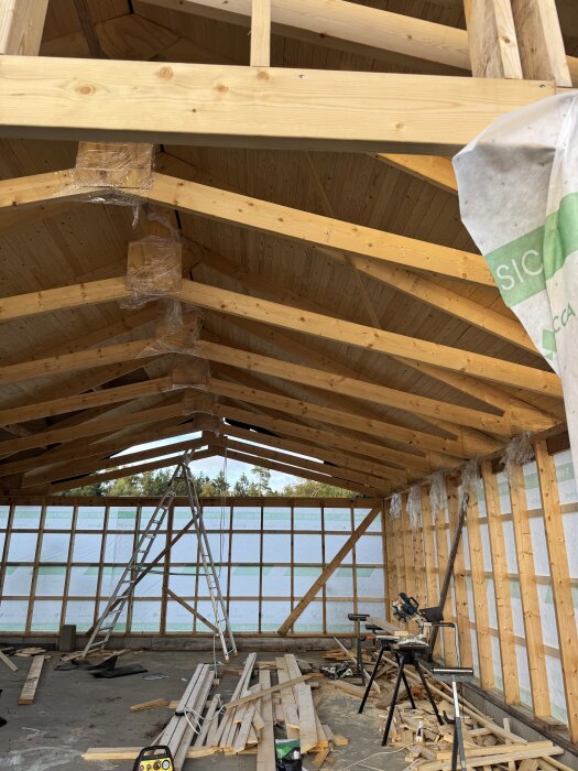 Bygge av garageinteriör med öppet tak, träbalkar, stege och byggmaterial utspritt på betonggolvet.