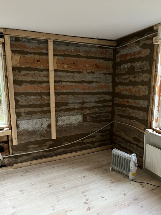 Vägg med trästommar och lerklining, kabel längs golvet och ett portabelt element på trägolv. Fönster syns delvis till höger.