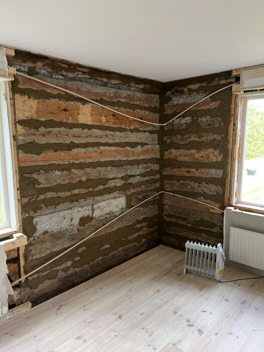 Vägg med synliga stockar och lerklinade skarvar, golv av ljust trä, element på golvet, två fönster med dagsljus som strömmar in.