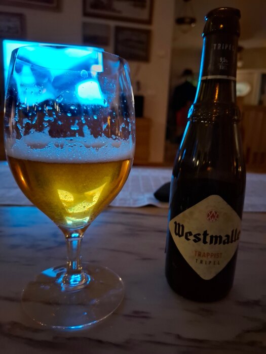 Flaska och glas med Westmalle Trappist Trippel öl på bord, blått sken i bakgrunden.