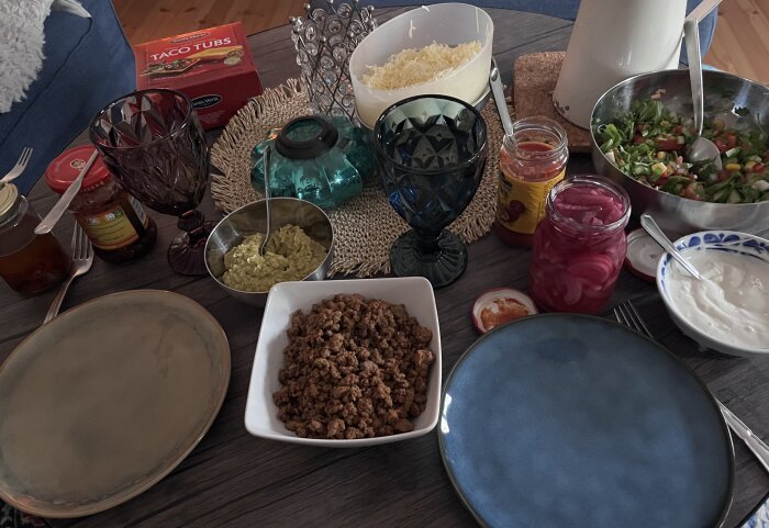 Tacomiddag med olika tillbehör på ett bord, inklusive köttfärs, guacamole, sallad, ost, picklad rödlök och gräddfil, samt tomma tallrikar.