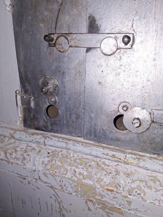 Närbild av en gammal kakelugnslucka i metall, med ventiler för luftflödesjustering. Vit färgflagor syns på omgivande murverk.