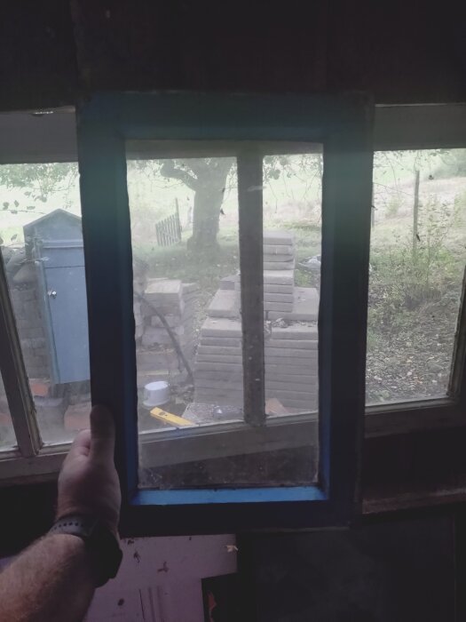 Fönsterram som hålls i handen vid ljusinsläppet på ett utedass, med utsikt över trädgård och träd genom smutsiga rutor.