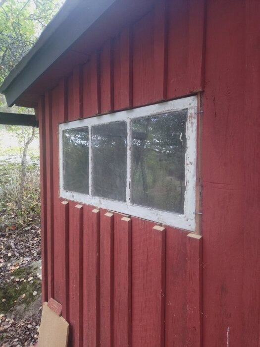 Rödmålat utedass med nytt ljusinsläpp, tredelat fönster och vit ram, omgivet av natur.