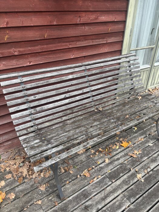 Sliten träbänk med lockläkt framför faluröd trävägg, omgivet av höstlöv på en altan av träplankor.